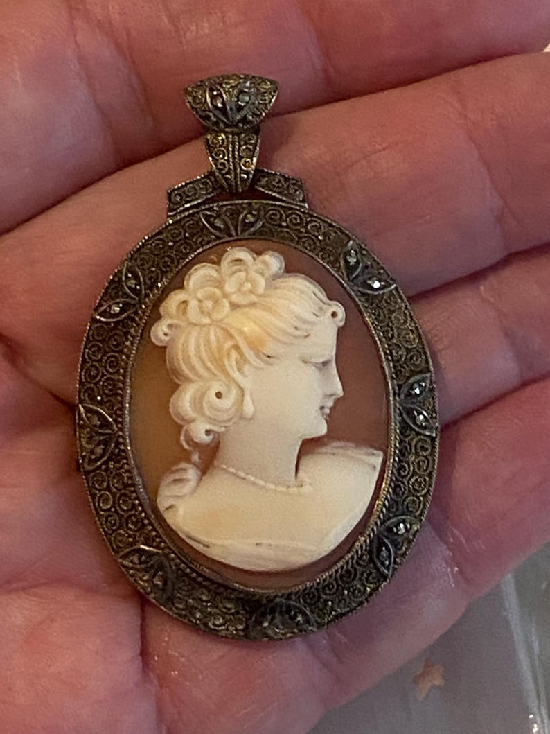 May include: Oval cameo pendant featuring a profile of a woman with a floral hairpiece. The cameo is set in a decorative silver-toned frame with marcasite accents. The pendant has a detailed bail.