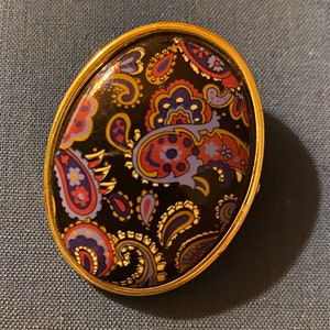May include: An oval-shaped brooch featuring a gold-toned metal frame surrounding a black insert decorated with a colorful paisley pattern in shades of red, blue, and yellow. The surface of the central insert is smooth and glossy.