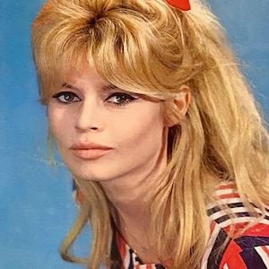 以下が含まれることがあります： 赤いリボンでスタイリングされたブロンドの髪の女性の肖像画。赤、白、黒のストライプのトップスを着ています。背景は無地の水色です。