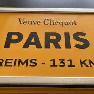 Puede incluir: Un letrero naranja con las palabras "Veuve Clicquot PARIS" en letras negras. Debajo, se lee "REIMS - 131 KM". El letrero tiene un borde plateado.