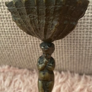 Puede incluir: Una escultura de bronce de una figura que sostiene un cuenco en la cabeza. La figura está de pie sobre un pedestal.