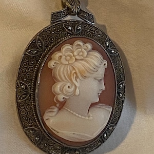 May include: Oval cameo pendant featuring a profile of a woman with a floral hairpiece and pearl necklace. The cameo is set in a silver-toned frame with decorative detailing and small accent stones. The pendant has a decorative bail.