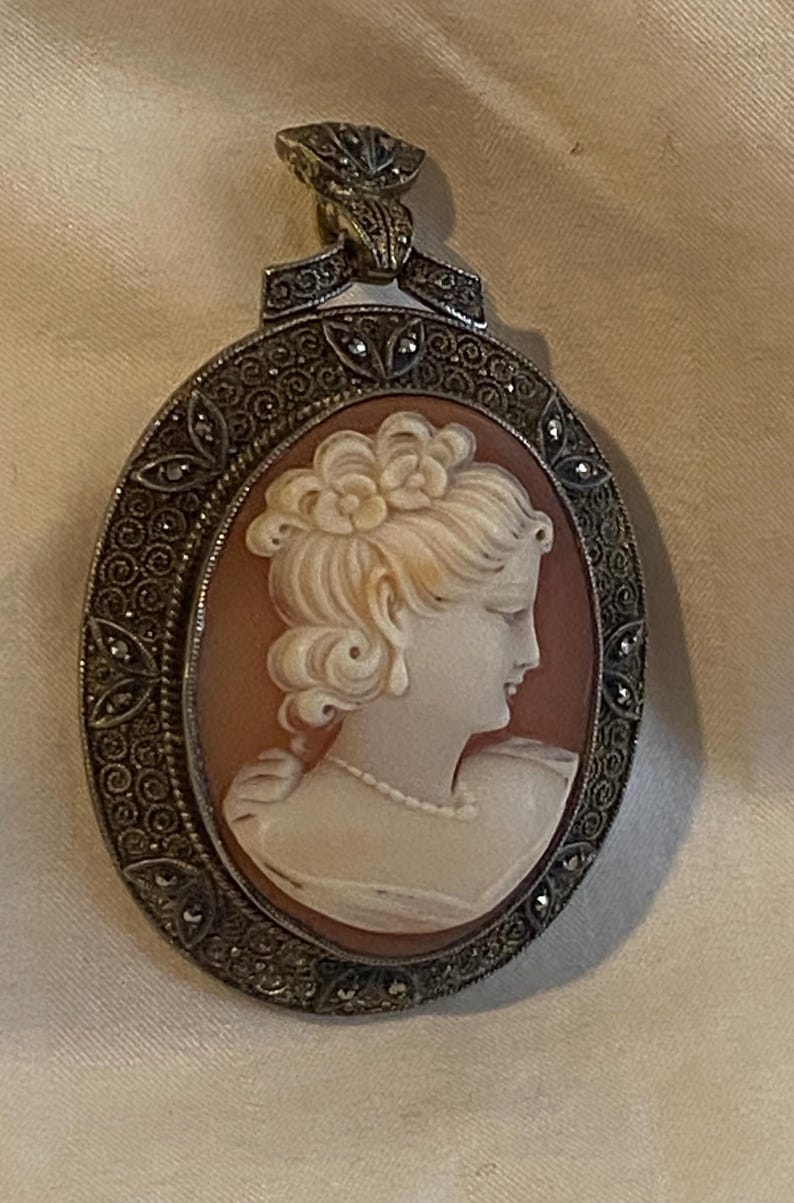 May include: Oval cameo pendant featuring a profile of a woman with a floral hairpiece and pearl necklace. The cameo is set in a silver-toned frame with decorative detailing and small accent stones. The pendant has a decorative bail.