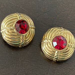 May include: Pair of gold-toned clip-on earrings. Each earring features a round, faceted, red gemstone set in a textured, circular frame with rope-like detailing. The earrings have a vintage aesthetic.
