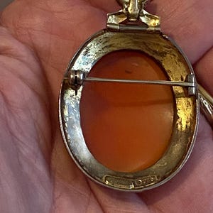 May include: A vintage oval cameo brooch with a gold-toned frame and a pin closure. The cameo features an orange-toned relief carving. The brooch has a decorative top element and is marked "STERLING".