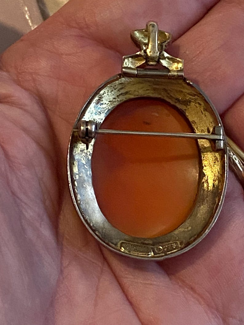 May include: A vintage oval cameo brooch with a gold-toned frame and a pin closure. The cameo features an orange-toned relief carving. The brooch has a decorative top element and is marked "STERLING".