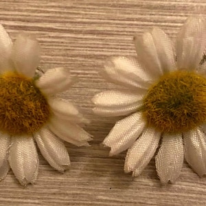 Puede incluir: Dos pinzas para el pelo con forma de flor de margarita. Cada pinza presenta pétalos blancos que rodean un centro amarillo. Las flores están sobre una superficie de madera marrón claro. Ideal para añadir un toque floral a cualquier peinado.