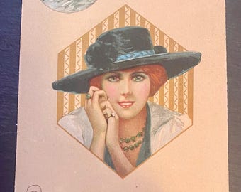 May include: A vintage postcard featuring a woman wearing a black hat with a blue ribbon and a green necklace. She is looking to the side and has her hand on her chin. A white cat is pictured in a circle in the upper left corner of the postcard.