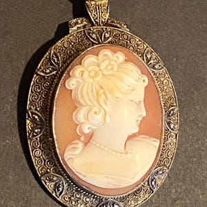 May include: Oval cameo pendant featuring a profile of a woman with a floral hairpiece and a pearl necklace. The cameo is set in a filigree frame with small, sparkling accents. The pendant has a decorative bail.