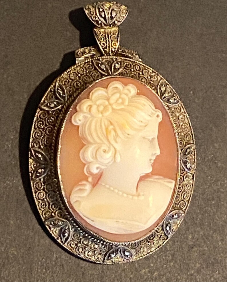 May include: Oval cameo pendant featuring a profile of a woman with a floral hairpiece and a pearl necklace. The cameo is set in a filigree frame with small, sparkling accents. The pendant has a decorative bail.