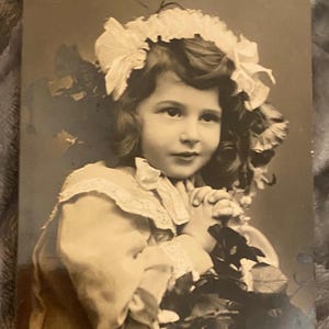 Può includere: Ritratto in seppia di una bambina, probabilmente una ragazza, con capelli ricci e un copricapo floreale. Indossa un vestito con finiture in pizzo e tiene dei fiori. L'immagine ha un'estetica vintage.