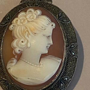 May include: Oval cameo pendant featuring a profile of a woman with a floral hairpiece and a pearl necklace. The cameo is set in a decorative silver-toned frame with intricate detailing and small embellishments.