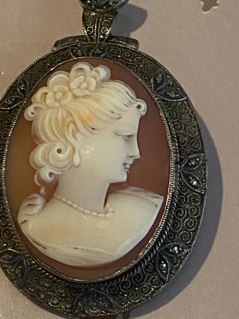 May include: Oval cameo pendant featuring a profile of a woman with a floral hairpiece and a pearl necklace. The cameo is set in a decorative silver-toned frame with intricate detailing and small embellishments.
