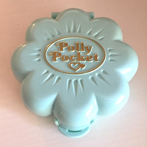 Può includere: Un compatto Polly Pocket azzurro a forma di fiore. Il compatto ha un ovale dorato al centro con le parole "Polly Pocket" e un cuore. Il compatto è chiuso e ha una piccola chiusura.