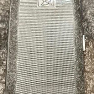 May include: Silver cigarette case with a textured, cross-hatched pattern. The case has ornate floral scrollwork around the edges and a small rectangular cartouche with the initials "H.L.".
