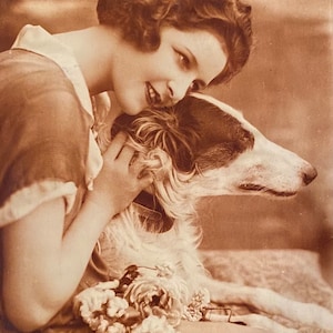Könnte beinhalten: Sepiafarbene Vintage-Fotografie einer Frau, die einen Borzoi-Hund umarmt. Der Hund hat ein langes, schlankes Gesicht mit weißem und braunem Fell. Die Frau trägt ein Kleid und hat kurzes, lockiges Haar. Ein Blumenstrauß liegt auf dem Stoff.
