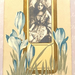 Peut inclure: Carte postale vintage avec un portrait en noir et blanc d'une femme en robe à fleurs, encadré d'une bordure dorée. La carte est ornée de fleurs de crocus bleues et blanches et de feuilles vertes. Texte manuscrit dans le coin inférieur droit.