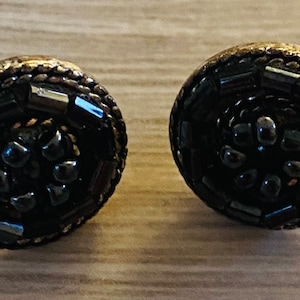 Puede incluir: Un par de pendientes de botón de estilo vintage. Cada pendiente presenta un diseño circular oscuro con un borde de cuentas y un grupo central de pequeñas cuentas oscuras. Los pendientes tienen un acabado metálico, posiblemente de latón.