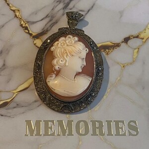 May include: Oval cameo pendant with a profile of a woman with a pearl necklace, set in a silver-toned frame. The pendant is on a marble surface with the word "MEMORIES" in gold lettering.
