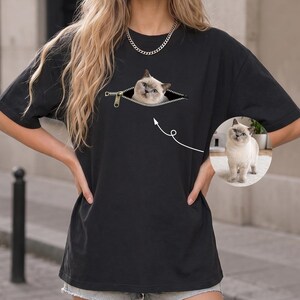 Op de afbeelding: Een zwart T-shirt met korte mouwen en een afbeelding van een rits die openstaat, waar een lichtgekleurde kat uit tevoorschijn komt. Een witte pijl wijst van de afbeelding naar een cirkelvormig kader met de kat in zijn geheel afgebeeld.