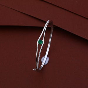 Peut inclure: Un bracelet jonc articulé de couleur argentée avec un design à double tige. La partie avant est ornée d'une pierre précieuse verte unique de taille ronde, entourée de deux rangées délicates de petites pierres claires serties pavé.