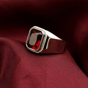 May include: A silver ring with a large, rectangular, deep red gemstone. The ring has a polished finish and is set against a burgundy background, highlighting the gemstone's color and shape. The ring's design is simple yet elegant.