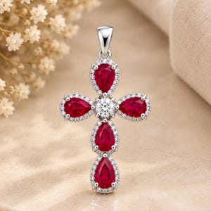 May include: A silver cross pendant featuring four pear-cut deep red rubies and a single round brilliant-cut clear stone at the center. Each ruby is surrounded by a halo of small white pavé stones, with a silver bail at the top for a chain.