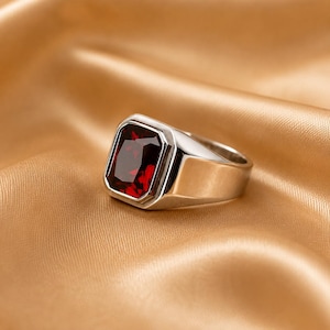 May include: A silver-toned ring featuring a large, rectangular, deep red faceted gemstone set in a smooth, polished metal band. The ring has a substantial, solid construction with a wide shank that tapers slightly towards the back of the band.