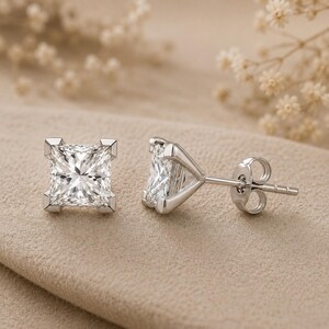 May include: A pair of silver-toned stud earrings featuring square-cut clear stones. One earring is shown from the front, displaying the faceted surface of the stone, while the other is shown from the side, revealing the metal setting and post back.