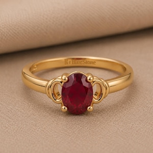May include: A gold ring featuring a central oval-cut deep red gemstone held by four prongs. The band has a smooth, polished finish and includes decorative scroll-like metalwork on either side of the stone. The interior band is engraved with BlueStone.