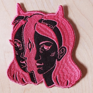 May include: Embroidered patch with a design of two faces in black and pink. The faces are side-by-side, with one having a third eye. The patch has a pink outline and hair, set against a light wood background.