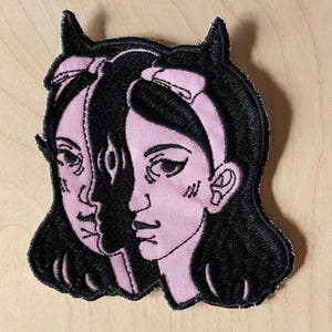 Może przedstawiać: Haftowana naszywka przedstawiająca stylizowaną ilustrację twarzy kobiety podzielonej na dwie części, ciemną i jasną. Projekt obejmuje czarne włosy, różową twarz i kokardę. Naszywka ma czarną obwódkę.