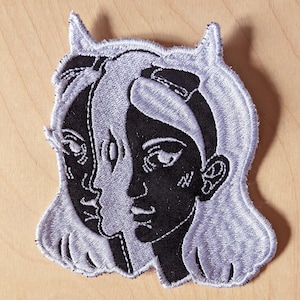 Op de afbeelding: Geborduurde patch met een gespleten gezichtsontwerp in zwart en grijs. Eén kant van het gezicht is zwart, de andere grijs, met een gedeeld oog en een hoornachtig detail aan de bovenkant. De patch heeft een gestikte rand.