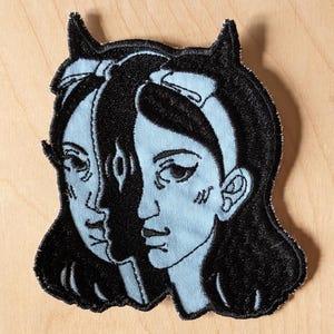 Könnte beinhalten: Gestickter Aufnäher mit einem geteilten Gesicht einer Frau mit schwarzen Haaren, Hörnern und einer Schleife. Das Gesicht auf der linken Seite ist hellblau, während die rechte Seite schwarz ist. Der Aufnäher hat einen schwarzen Rand und liegt auf einer hellen Holzoberfläche.