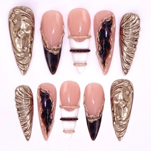 May include: A set of ten artificial nails in various designs. Some nails are gold-colored with embossed patterns, while others feature a nude base with gold accents and dark brown tips. One nail has a white and black design.