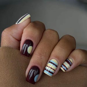 May include: Manicured fingernails featuring a mix of designs. Some nails display horizontal stripes in pale yellow, light blue, and dark brown, while others are painted solid dark brown with a single pale yellow or light blue circular accent.