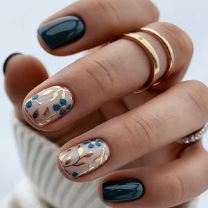 May include: Manicured nails featuring a combination of solid dark teal polish and a light nude base with metallic gold leaf foliage and small blue floral accents. Two plain gold bands are worn on the middle finger, and a silver ring is visible.