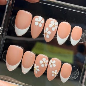 May include: A set of artificial nails in a clear plastic case. The nails feature a peach-colored base with white tips and white flower designs with gold accents. Some nails have a French tip design.