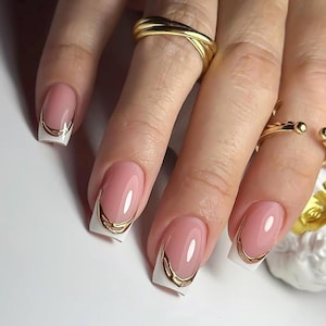 May include: Close-up of manicured nails with a pink base, white tips, and gold metallic accents. The nails are oval-shaped and feature a French manicure design with a modern twist. Several gold rings adorn the fingers.