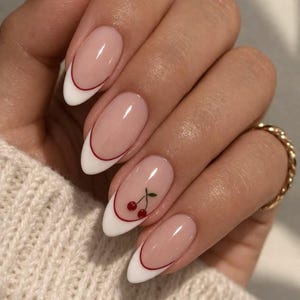 May include: Almond-shaped nails featuring a classic French manicure design with a nude base and crisp white tips, accented by a thin red line separating the colors. One nail displays a small, detailed graphic of two red cherries with green stems.
