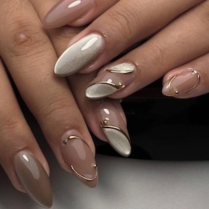 May include: Close-up of a hand with almond-shaped, professionally manicured nails. The nails feature a variety of designs, including solid neutral colors, metallic silver, and gold accents. Some nails have gold wire and small gold bead details.