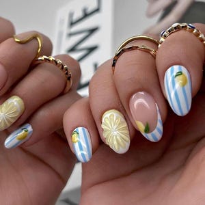 May include: Oval-shaped fingernails featuring a mix of light blue and white vertical stripes, yellow lemon illustrations, and three-dimensional pale yellow flower accents. The nails are adorned with gold-toned metal rings on the fingers.