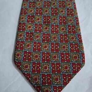 Puede incluir: Una corbata roja con un patrón cuadrado repetido. Cada cuadrado presenta un diseño dorado, verde y blanco sobre un fondo rojo. La corbata está hecha de una tela suave y tiene un extremo puntiagudo clásico.