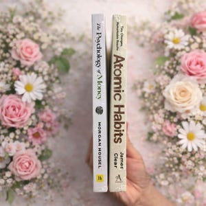 May include: Two hardcover books, "The Psychology of Money" and "Atomic Habits," are displayed vertically. The first book has a white cover with black text, and the second has a cream cover with brown text. The background features pink roses and white daisies.