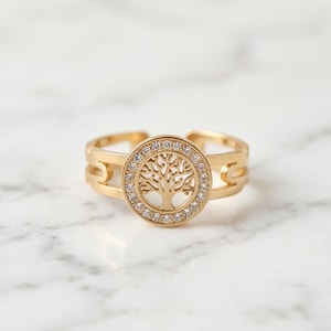 Puede incluir: Anillo dorado con un diseño de árbol de la vida. El árbol está rodeado de pequeñas piedras preciosas transparentes. El anillo tiene un diseño de caña dividida.