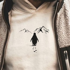 May include: Cream-colored hoodie with a black penguin graphic walking towards mountains. The hoodie is worn under a beige fleece jacket with brown accents and a zippered pocket. The image has a blurred outdoor background.