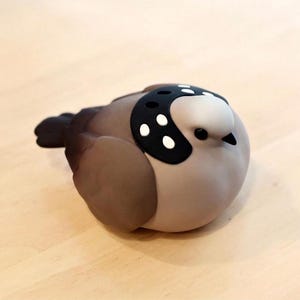 Puede incluir: Figura de pájaro de cerámica en tonos marrón, gris y blanco. El pájaro tiene un parche negro con puntos blancos en la cabeza. La figura descansa sobre una superficie de madera de color claro.
