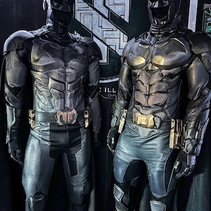 May include: Two Batman costumes are displayed, showcasing intricate details. The suits are predominantly dark gray and black, with sculpted chest plates, utility belts, and helmets. The costumes are designed with a focus on a muscular physique, and the overall aesthetic is sleek and modern.
