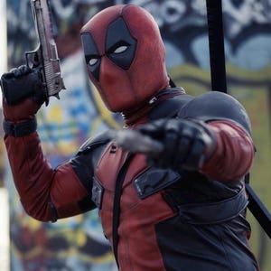 May include: A person in a Deadpool costume, holding a silver handgun and a blade. The costume is red and black with a mask featuring white eye shapes. The background shows graffiti art.