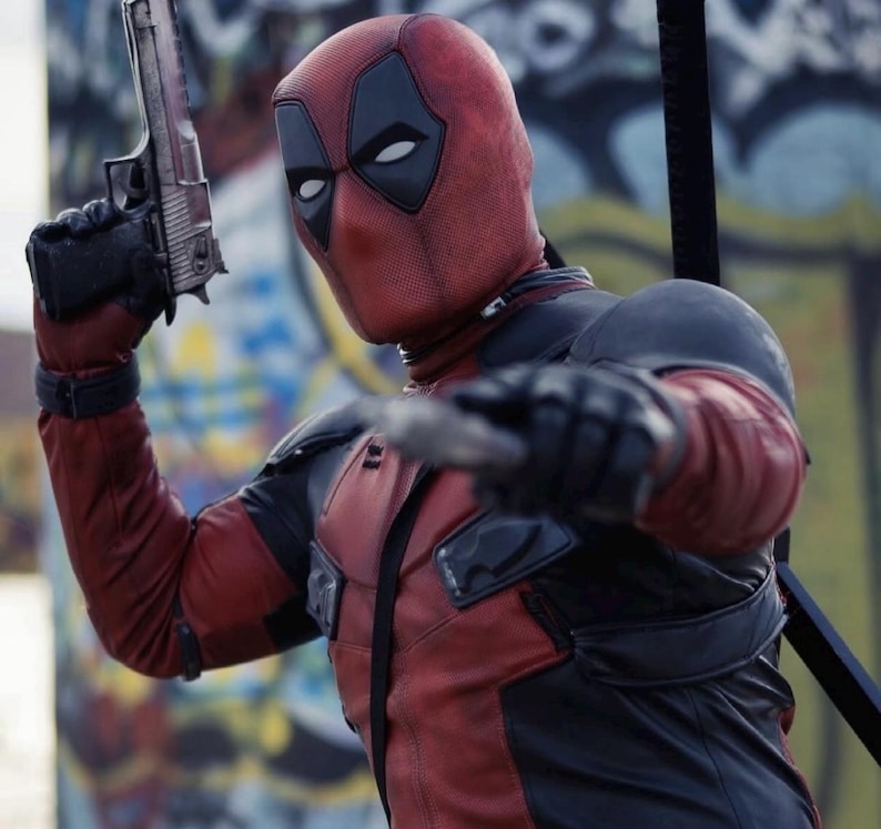 May include: A person in a Deadpool costume, holding a silver handgun and a blade. The costume is red and black with a mask featuring white eye shapes. The background shows graffiti art.